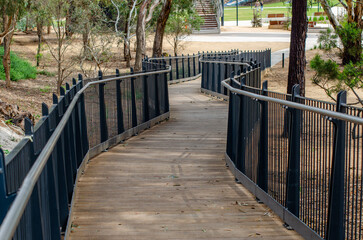 A curved wooden boardwalk with metal safety railings winds through an Australian park, providing an accessible route through public parkland—an inclusive community amenity.