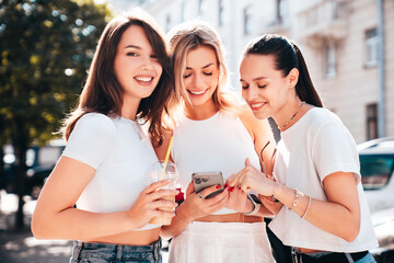 Three young beautiful smiling trendy female. Carefree women posing in street. Positive models having fun. Cheerful and happy. Hold smartphone, look at mobile cellphone screen, use phone apps
