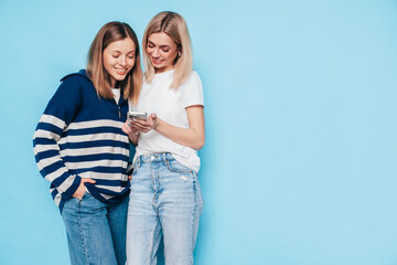 Two young beautiful smiling brunette hipster female in trendy summer clothes. Carefree women posing near blue wall. Positive models having fun. Cheerful and happy. Holding smartphone. Using apps