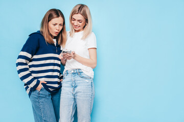 Two young beautiful smiling brunette hipster female in trendy summer clothes. Carefree women posing near blue wall. Positive models having fun. Cheerful and happy. Holding smartphone. Using apps