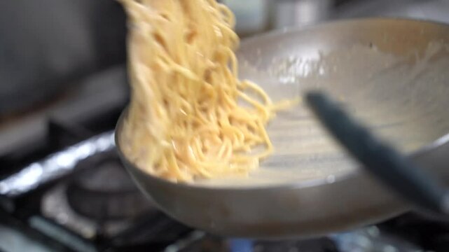A close-up of creamy spaghetti being prepared in a large pan on a stovetop. The rich sauce coats the pasta evenly, creating a smooth and appetizing texture.
