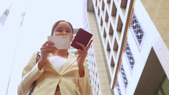 Asian female tourist checks passport validity in front of city office building and checks air ticket in preparation for travel.