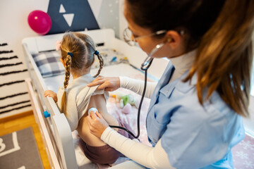 Rear view of pediatrician with stethoscope examining little girl's lungs at home checkup.
