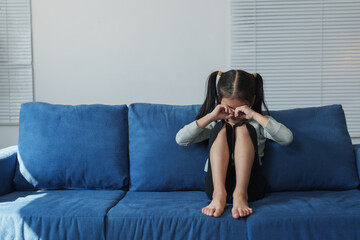 Young girl with pigtails sits alone on a blue couch, covering her eyes with her hands. The room is...