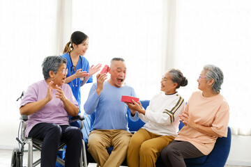 Fototapeta premium Group of happy senior friends, including a woman in a wheelchair, share joyful moment while exchanging gifts in bright living room. A nurse in scrubs claps along, caregiver, celebration, companionship