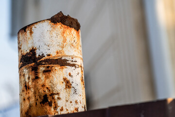 A close-up of a rusted metal pipe with heavy corrosion and weathering. The rust forms an interesting texture with a mix of brown, orange, and white hues. The background is blurred