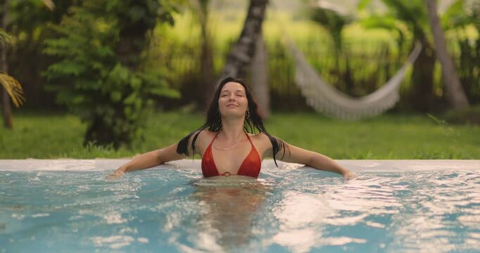A tranquil woman in a red bikini relaxes in a pool with her arms spread, enjoying the peaceful ambiance of a lush, tropical backdrop. Perfect for themes of luxury, wellness, and vacation.