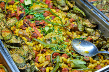 A vibrant dish of cooked beans, sausage, zucchini, and peppers, garnished with fresh herbs, served in a stainless steel tray with a spoon.