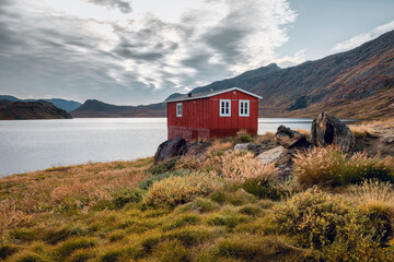 Red wooden cottage on shore of Innajuattoq lake on Arctic Circle Trail during a day in southwest...