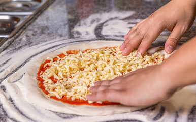 Closeup of man standing at board and sprinkling cheese on pizza dough with tomato sauce. Man adding cheese to pizza at table, closeup. Cook in the kitchen putting the ingredients on the pizza