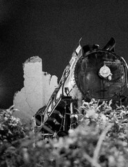  A rusted steam locomotive stands in front of a crumbling wall at night, bathed in golden light. The eerie scene evokes nostalgia and mystery, framed by blurred autumn foliage.