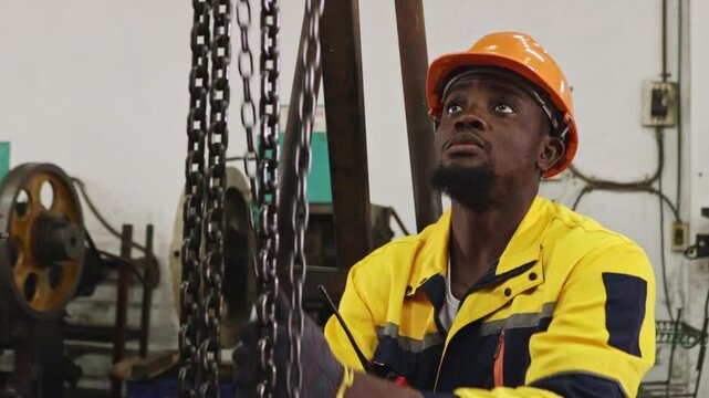 African American factory worker wearing hard hat and gloves pulls chain on steel beam for safety, while lifting and moving heavy machinery placed on floor in automotive parts lathe workshop.