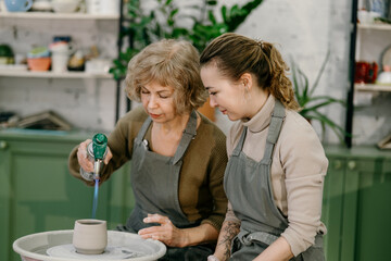 Hands-on learning, senior woman mastering pottery techniques using rotating wheel Mentor teaches pottery fundamentals, demonstrating sculpting skills on an electric wheel