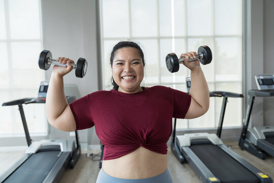 Happy smiling Asian young plus size woman lifting dumbbell weight in fitness club. Young overweight woman holding dumbbell workout at gym. Asian woman exercise training with weight, healthy lifestyle