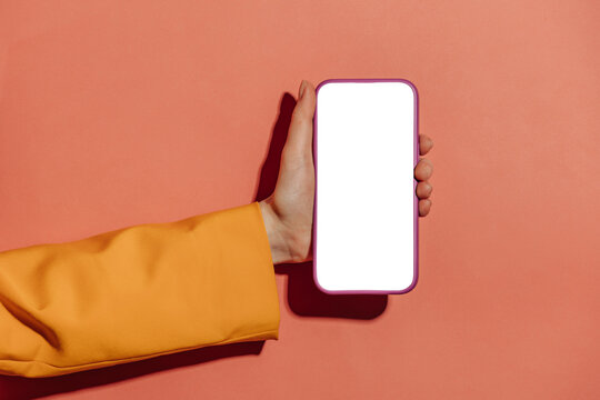 woman's hand in a yellow blazer holds a mobile phone with a white screen mockup against a pink background