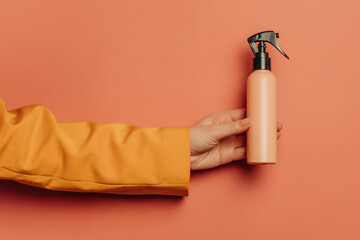 woman's hand in a yellow blazer holds a tanning spray bottle against a pink background