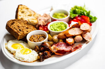 A delicious breakfast platter featuring fried eggs, crispy bacon, sausages, fresh vegetables, assorted dips, and slices of bread, served on a white oval plate. Perfect for a hearty morning meal.