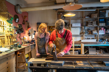 Novice woman learns and observes master carpenter working on machine in workshop. Husband and wife work together, manual family run furniture production. Hands on learning, teamwork, small business