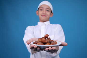 Portrait Of Muslim Teenage Boy Offering Dates Fruit To Front Over Blue Background 