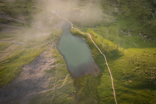 Aerial view of tranquil Lac Vert surrounded by serene mountains and a winding road with a cottage, Val d'Illiez, Switzerland.