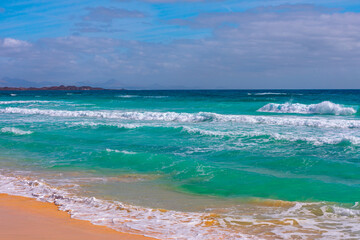 Scenic view of a beach with turquoise waves crashing onto sandy shore under partly cloudy sky with distant mountains visible on horizon. Coastal scene featuring tranquil beach with emerald green waves