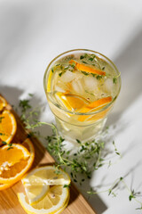 ice tea with citrus fruits and thyme in a glass on white background. iced tea with lemon and orange in drinking glass with fruits on cutting board with towel on side. harsh lighting with shadows.