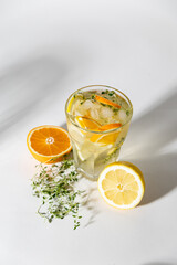 ice tea with citrus fruits and thyme in a glass on white background. iced tea with lemon and orange in drinking glass with fruits on cutting board with towel on side. harsh lighting with shadows.