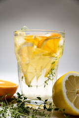 ice tea with citrus fruits and thyme in a glass on white background. iced tea with lemon and orange in drinking glass with fruits on cutting board with towel on side. harsh lighting with shadows.