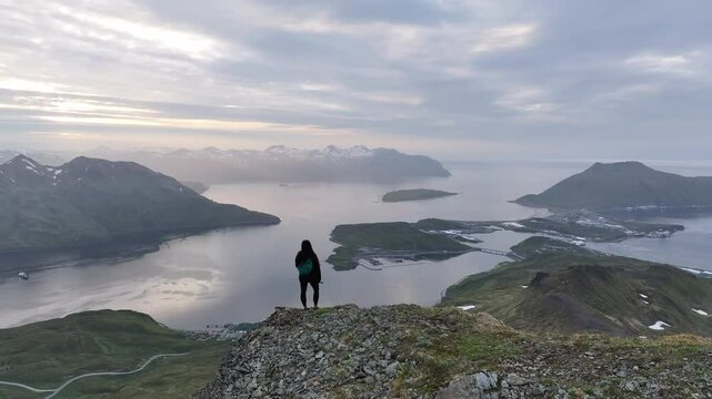 Aerial view of a woman standing on a rock overlooking a serene sunset at Captains Bay with rugged mountains and tranquil water, Unalaska, United States.