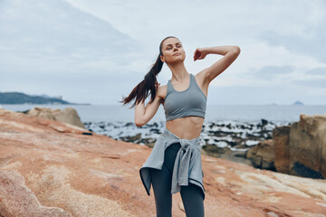 Fit young woman stretching on rocky beach in athletic wear, showcasing healthy lifestyle and fitness, ocean background, cloudy weather, and serene atmosphere