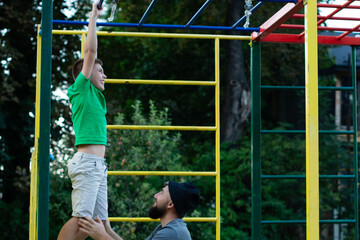 A young boy performs pull-ups, his father supporting him from below. The scene unfolds on a vibrant climbing frame set against a backdrop of lush greenery.