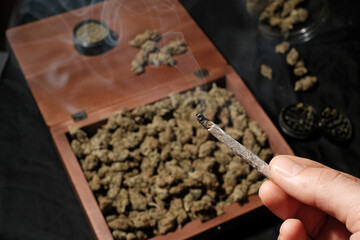 Smoking medical marijuana joint in a man's hand on a background of cannabis buds, grinder and jars with cones