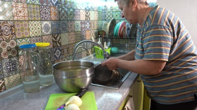 An elderly woman washes a pot in the kitchen sink. Home work