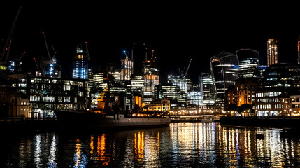 Obraz premium Night Cityscape, Illuminated Skyscrapers Reflecting on Water, a Vessel Docked, Construction Cranes Visible in the Background. City at Night, Urban Architecture, Modern Buildings.