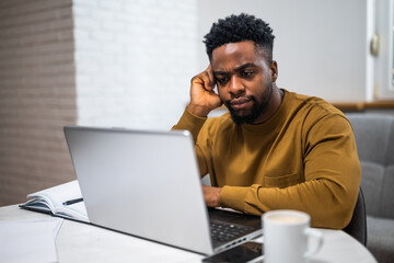 Overworked and anxious black freelancer using laptop while working remotely at his home office.