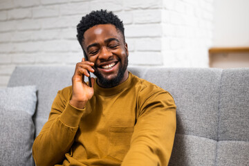 Happy African American man smiling and enjoying talking on the mobile phone while relaxing on sofa at his home