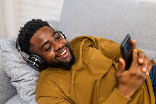 Cheerful young  black man enjoying using wireless headphones and mobile phone while lying on sofa in his cozy home.