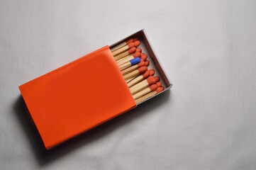 A lone blue matchstick pops among red ones in an open orange matchbox against a white backdrop.