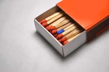 A single blue match head stands out among a bunch of red matches inside an orange matchbox.