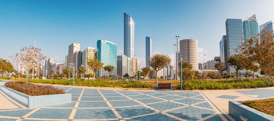Obraz premium Geometric blue and white tiled pavement and green areas in Abu Dhabi Corniche public park under a clear blue sky with modern office buildings