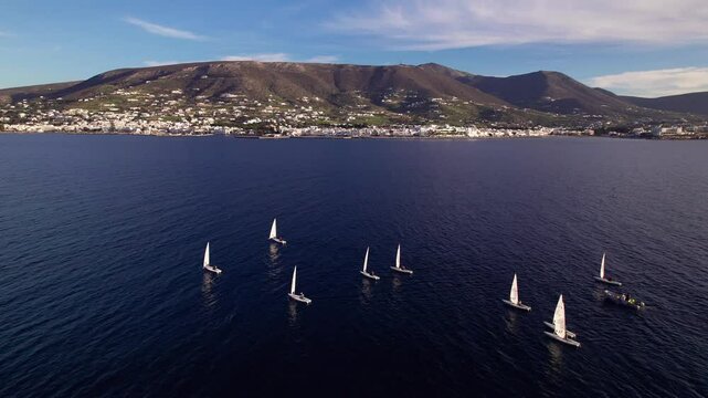 Drone shot of sail&iota;ng boats with sails outside of Parikia town on Paros island in Greece. Drone flying towards them showing Parikia town behind.