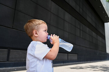 A little boy dressed in a simple white shirt takes a break from his adventures to sip water from a...