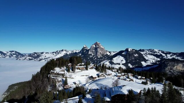 An iconic view of the Mythen mountains towering over the picturesque village of Stoos. Snow-covered rooftops and alpine scenery create a stunning winter postcard
