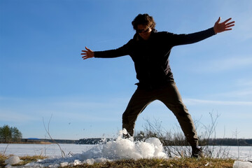 Playful man breaks a block of ice near frozen river on nature in sunny day. Guy throwing ice on the land to break it up. Large piece of ice breaks up into small fragments. Winter, early spring season.