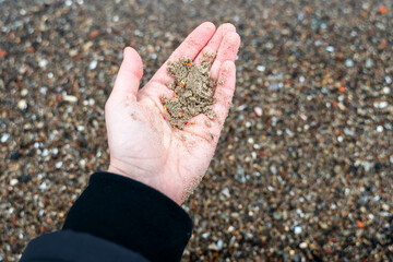 Open palm holds sand mixed with tiny microplastic particles against sandy sea shore, highlighting environmental pollution in cold weather