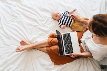 Mother working at laptop playing with baby in bed at home.