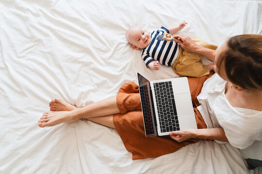 Mother working at laptop playing with baby in bed at home.