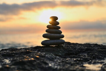 A carefully balanced tower of smooth stones stands majestically against a stunning sunset. The vibrant colors of dusk reflect off the calm water, enhancing the tranquil atmosphere.