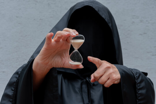 A figure in a black hooded cloak holds an hourglass on a gray background.