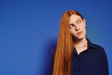 Young queer man with long red hair showcases stylish fashion against a vibrant blue backdrop